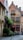 A quaint narrow street lined with old brick buildings, including a charming establishment with red-framed windows and a sign reading 'Vlaamsche Pot.' The façade is adorned with greenery and flowers. The cobblestone path and cozy outdoor seating evoke a sense of vintage European architecture.