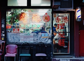 A storefront of a traditional barbershop with colorful neon signs and Chinese characters. Vintage chairs are placed outside, and a 'No Smoking' sign is visible on the window. Inside, a person can be seen getting a haircut.