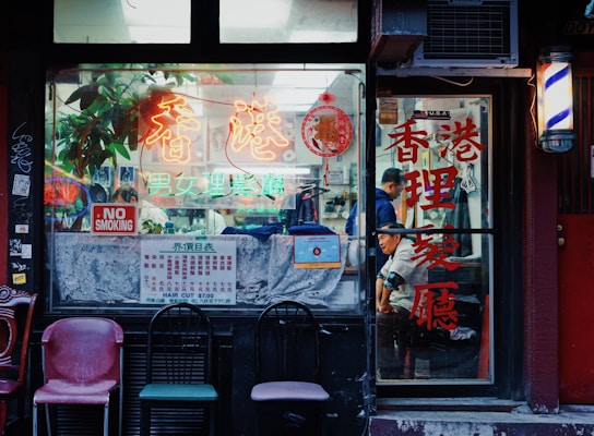 A storefront of a traditional barbershop with colorful neon signs and Chinese characters. Vintage chairs are placed outside, and a 'No Smoking' sign is visible on the window. Inside, a person can be seen getting a haircut.