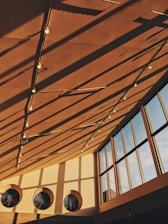 Warm sunlight casts long shadows from a geometric ceiling structure with hanging lights. Large circular vents and tall glass windows contribute to the modern architectural ambiance.