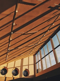 Warm sunlight casts long shadows from a geometric ceiling structure with hanging lights. Large circular vents and tall glass windows contribute to the modern architectural ambiance.