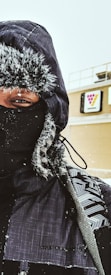 A person dressed in heavy winter clothing including a hooded jacket and face mask, with snowflakes visible on their clothing. In the background, there is a building with what appears to be a Seven-Eleven logo.