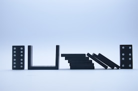 A series of black domino pieces placed in a row against a light blue background. Some of the dominoes are standing upright, while others are in the process of toppling over, demonstrating a chain reaction.