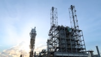 An industrial oil refinery structure is set against a backdrop of a clear blue sky. The facility features tall, intricate metal frameworks and towers, with the sun partially obscured behind one of the structures, creating a glow.