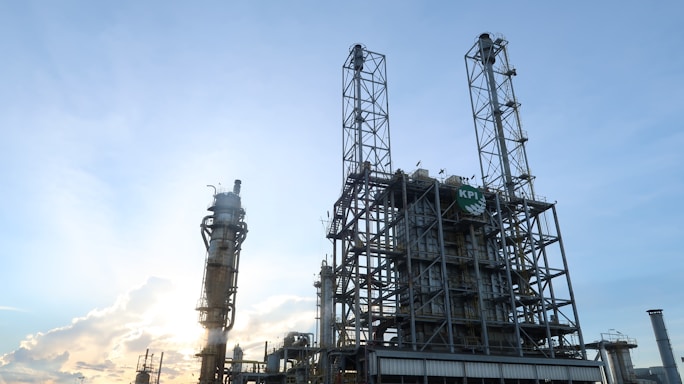 An industrial oil refinery structure is set against a backdrop of a clear blue sky. The facility features tall, intricate metal frameworks and towers, with the sun partially obscured behind one of the structures, creating a glow.