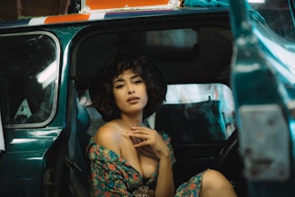 A person with curly hair is sitting in a vehicle. The individual is wearing a floral dress and is captured in a relaxed, confident pose. The interior of the car is visible, with details like upholstery and windows. The lighting emphasizes a chic, vintage ambiance.