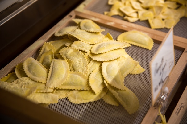 A wooden tray filled with uncooked ravioli pieces, each with a decorative edge, placed on a mesh surface for drying. The lighting highlights the texture and flour dusting on the pasta.