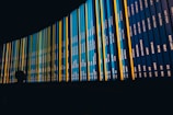A large curved display board shows rows of information with various city names and dates. The board is predominantly dark blue with bright yellow and teal strips. A silhouette of a person is visible in the foreground, observing the display.