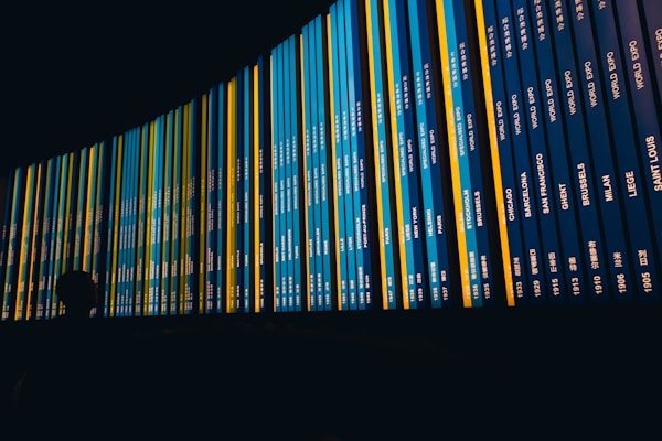 A large curved display board shows rows of information with various city names and dates. The board is predominantly dark blue with bright yellow and teal strips. A silhouette of a person is visible in the foreground, observing the display.