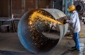A worker wearing a hard hat and protective gear uses an angle grinder to cut through a large metal pipe. Bright sparks are emitted from the cutting process, creating a vivid contrast against the industrial setting. In the background, there are various tools and equipment characteristic of a workshop environment.
