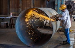 A worker wearing a hard hat and protective gear uses an angle grinder to cut through a large metal pipe. Bright sparks are emitted from the cutting process, creating a vivid contrast against the industrial setting. In the background, there are various tools and equipment characteristic of a workshop environment.