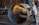 A worker wearing a hard hat and protective gear uses an angle grinder to cut through a large metal pipe. Bright sparks are emitted from the cutting process, creating a vivid contrast against the industrial setting. In the background, there are various tools and equipment characteristic of a workshop environment.