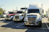 Several large semi-trucks parked in a row in a parking lot under a clear sky. The trucks are of varying colors, mostly white and red, and are positioned closely together. The area is well-lit by daylight, and there are some trees and other vehicles visible in the background.