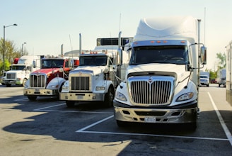 Several large semi-trucks parked in a row in a parking lot under a clear sky. The trucks are of varying colors, mostly white and red, and are positioned closely together. The area is well-lit by daylight, and there are some trees and other vehicles visible in the background.