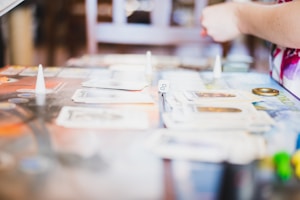 A board game is being played with various cards laid out on a table. Small cone-shaped markers and a die are visible. A person is interacting with the game, with their arm partially in view, suggesting active participation.