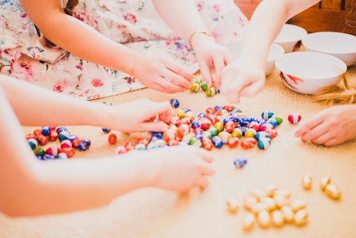 Several hands reaching for colorful assorted candy scattered on a beige surface, accompanied by empty white bowls with red patterns. A floral-patterned dress is partially visible.