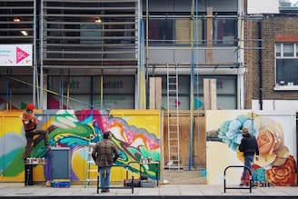 Three people are actively painting vibrant graffiti murals on a construction site barrier. The artwork is colorful and dynamic, featuring abstract shapes and floral designs. One person is perched on a stool, while another stands on the ground, both focused on their creative work. Ladders and various paint cans are scattered around the area.