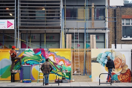 Three people are actively painting vibrant graffiti murals on a construction site barrier. The artwork is colorful and dynamic, featuring abstract shapes and floral designs. One person is perched on a stool, while another stands on the ground, both focused on their creative work. Ladders and various paint cans are scattered around the area.