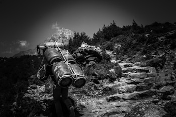 A person is hiking up a rocky mountain trail, carrying multiple gas canisters tied together. The path is steep and surrounded by dense vegetation, while a towering mountain peak looms in the background under a cloudy sky.