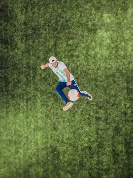 A person is performing a dynamic action with a soccer ball on a grassy field, wearing a striped jersey and a cap. The image is captured from above, highlighting the vibrant details of the grass and the focus on the movement.
