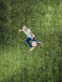 A person is performing a dynamic action with a soccer ball on a grassy field, wearing a striped jersey and a cap. The image is captured from above, highlighting the vibrant details of the grass and the focus on the movement.