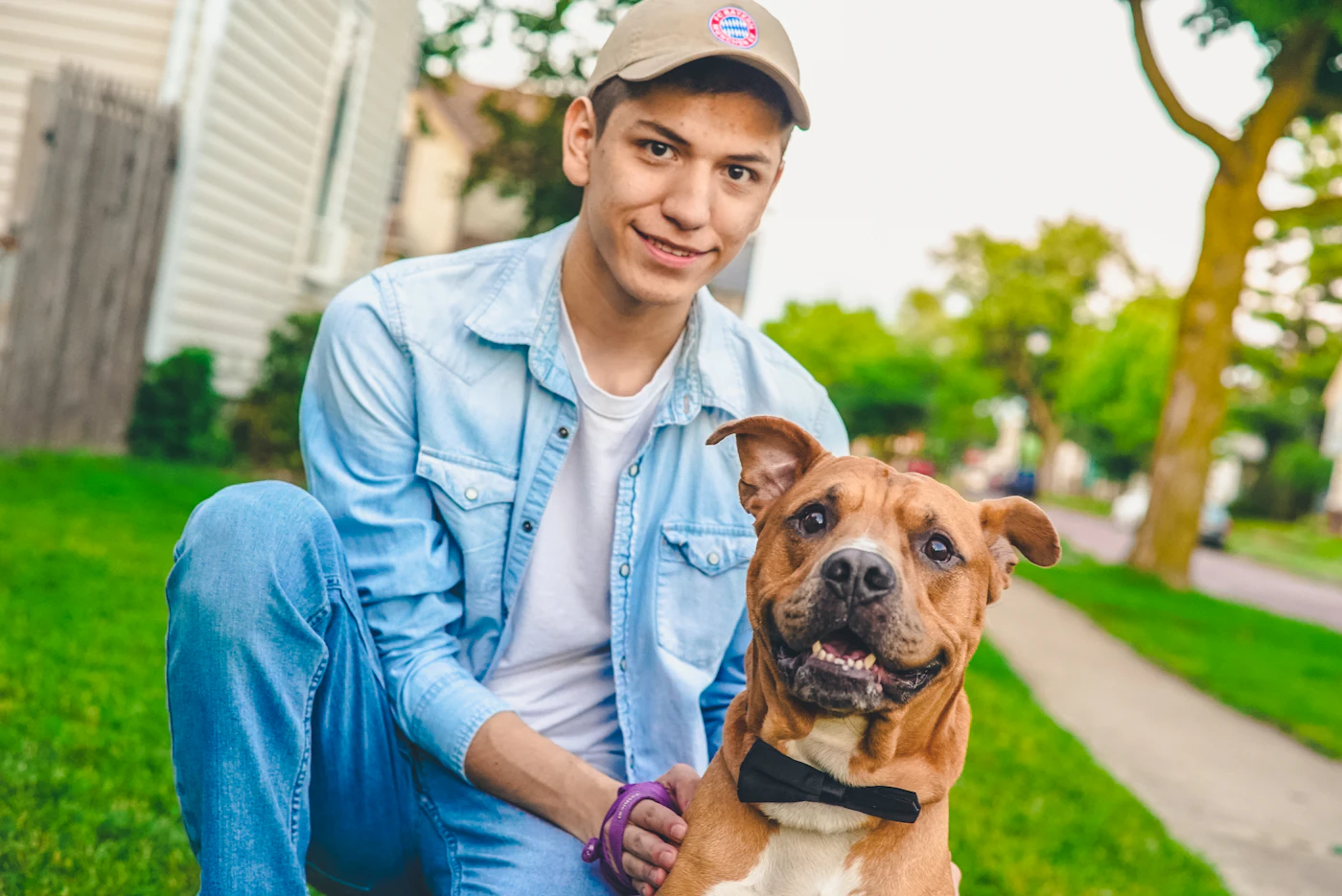 A young man in a denim shirt kneeling beside his calm dog on a tree-lined residential street