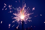 A sparkling firework emits bright sparks and subtle trails of light against a dark background. The central flare is intense and luminous, surrounded by smaller, scattered sparks that vary in color and intensity.