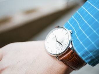 A polished wristwatch with a white dial and silver case is fastened to a brown leather strap. It is worn on the wrist of a person dressed in a blue checkered shirt.