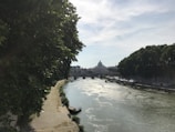 A serene river scene with lush green trees on both banks. A distant view of an iconic domed building appears through a bridge framing the sky. Rafts and tents line the water's edge under a partially cloudy blue sky.