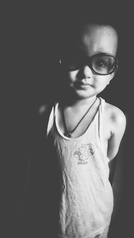 A young child wearing large sunglasses stands in soft lighting. They are wearing a sleeveless shirt with a playful graphic of a squirrel and the word 'SMART'. The image is in black and white, enhancing the contrast and shadows.