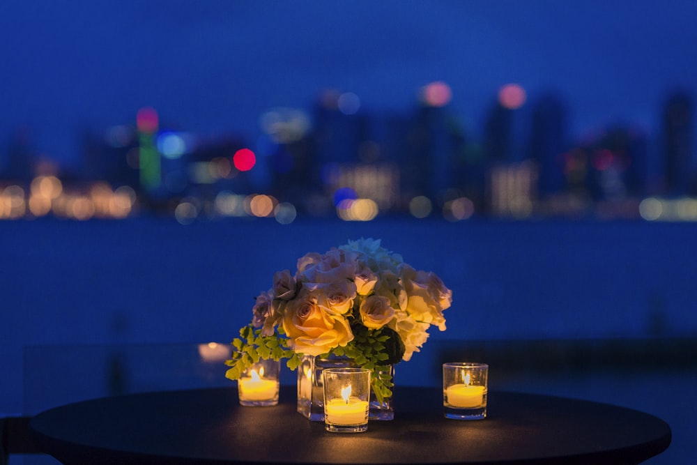 花瓶に白いバラの花束、茶色の丸い木製テーブルにティーライトキャンドルの奉納品が3つ
