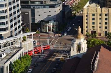 An urban scene with a prominent red tram traveling along a street, surrounded by modern buildings and a historical structure with a dome. Pedestrians are walking on the sidewalks, and there are several parked cars. Trees add greenery to the scene, while bright sunlight casts shadows.