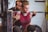 A woman is intensely focused on lifting a heavy barbell while a man stands closely behind her for support. She is wearing a maroon shirt and black knee sleeves, with visible tattoos on her arms and neck. The man behind her appears attentive, ensuring her safety during the lift.