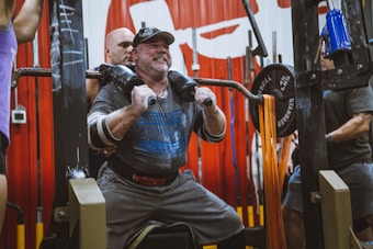 A man is performing a squat exercise with a barbell on his shoulders, assisted by a spotter. He is wearing a weightlifting belt and appears to be exerting significant effort. The gym setting includes weightlifting equipment and other people engaged in exercising.