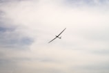 A sleek glider aircraft is silhouetted against a softly clouded sky, gracefully soaring to the right.