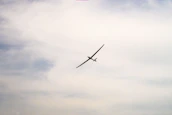 A sleek glider plane soaring silently over rolling hills under a bright sun.