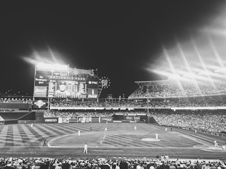 A lively baseball game at a large stadium under bright lights at night. The field is surrounded by a full audience, and the scoreboard displays various advertisements and player information. The atmosphere is energetic, with players visible on the field and a clear night sky above.