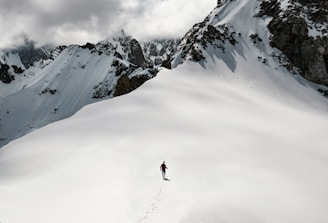A lone person is trekking through a vast expanse of snow in a mountainous area. The individual is leaving a trail of footprints behind and is surrounded by high, jagged peaks partially covered in snow. Above the mountains, heavy, cloudy skies dominate the scene.