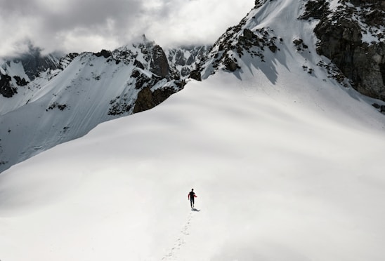 A lone person is trekking through a vast expanse of snow in a mountainous area. The individual is leaving a trail of footprints behind and is surrounded by high, jagged peaks partially covered in snow. Above the mountains, heavy, cloudy skies dominate the scene.