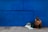 A man sits on the ground leaning against a large blue wall, appearing contemplative or weary. Beside him are a couple of bags, one of which is labeled 'homeless'. The wall and pavement create a stark background.