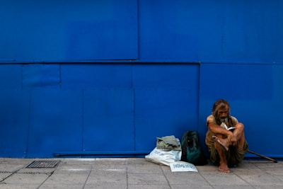 A man sits on the ground leaning against a large blue wall, appearing contemplative or weary. Beside him are a couple of bags, one of which is labeled 'homeless'. The wall and pavement create a stark background.
