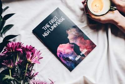 A book titled 'The First New Universe' lies on a white surface, featuring a cover with silhouettes against a cosmic backdrop. Nearby, pink flowers rest elegantly to the left while a pair of hands holds a lit candle in a cup on the right.