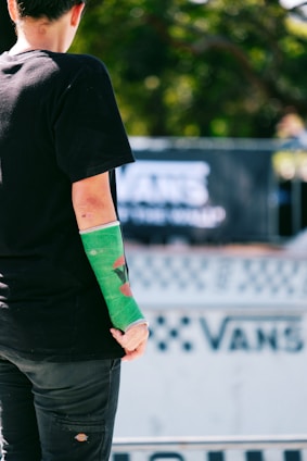 A person with a green cast on their arm is standing outdoors. The background displays a blurred event banner with a Vans logo, suggesting a skateboarding or sports-related event. The person wears a black t-shirt and pants, and there is a focus on their injured arm.