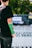 A person with a green cast on their arm is standing outdoors. The background displays a blurred event banner with a Vans logo, suggesting a skateboarding or sports-related event. The person wears a black t-shirt and pants, and there is a focus on their injured arm.