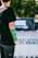 A person with a green cast on their arm is standing outdoors. The background displays a blurred event banner with a Vans logo, suggesting a skateboarding or sports-related event. The person wears a black t-shirt and pants, and there is a focus on their injured arm.