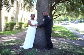 A couple is standing outdoors on either side of a large tree. The woman is wearing a white dress and holding a bouquet, while the man is dressed in a black suit. They both appear to be smiling and holding hands around the tree. The background features a building with ivy-covered walls and a lawn with trees and bushes under bright sunlight.