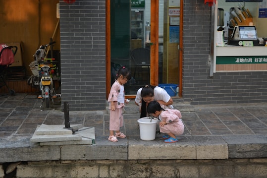 Three children are gathered around a white bucket on the pavement. Two of them are closely examining the bucket's contents, showing intense curiosity. The setting is outside a shop with dark brick walls and glass doors. There are bicycles parked nearby, and various shop items are visible through the shop window.