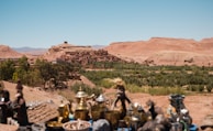 A historic fortified village or ksar nestled amidst a desert landscape, with clay buildings harmonizing with the surrounding reddish-brown mountains. Greenery, possibly palm groves, contrasts with the arid terrain. In the foreground, traditional artifacts and pottery are displayed on a table.