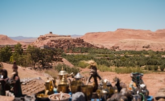 A historic fortified village or ksar nestled amidst a desert landscape, with clay buildings harmonizing with the surrounding reddish-brown mountains. Greenery, possibly palm groves, contrasts with the arid terrain. In the foreground, traditional artifacts and pottery are displayed on a table.