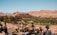A historic fortified village or ksar nestled amidst a desert landscape, with clay buildings harmonizing with the surrounding reddish-brown mountains. Greenery, possibly palm groves, contrasts with the arid terrain. In the foreground, traditional artifacts and pottery are displayed on a table.